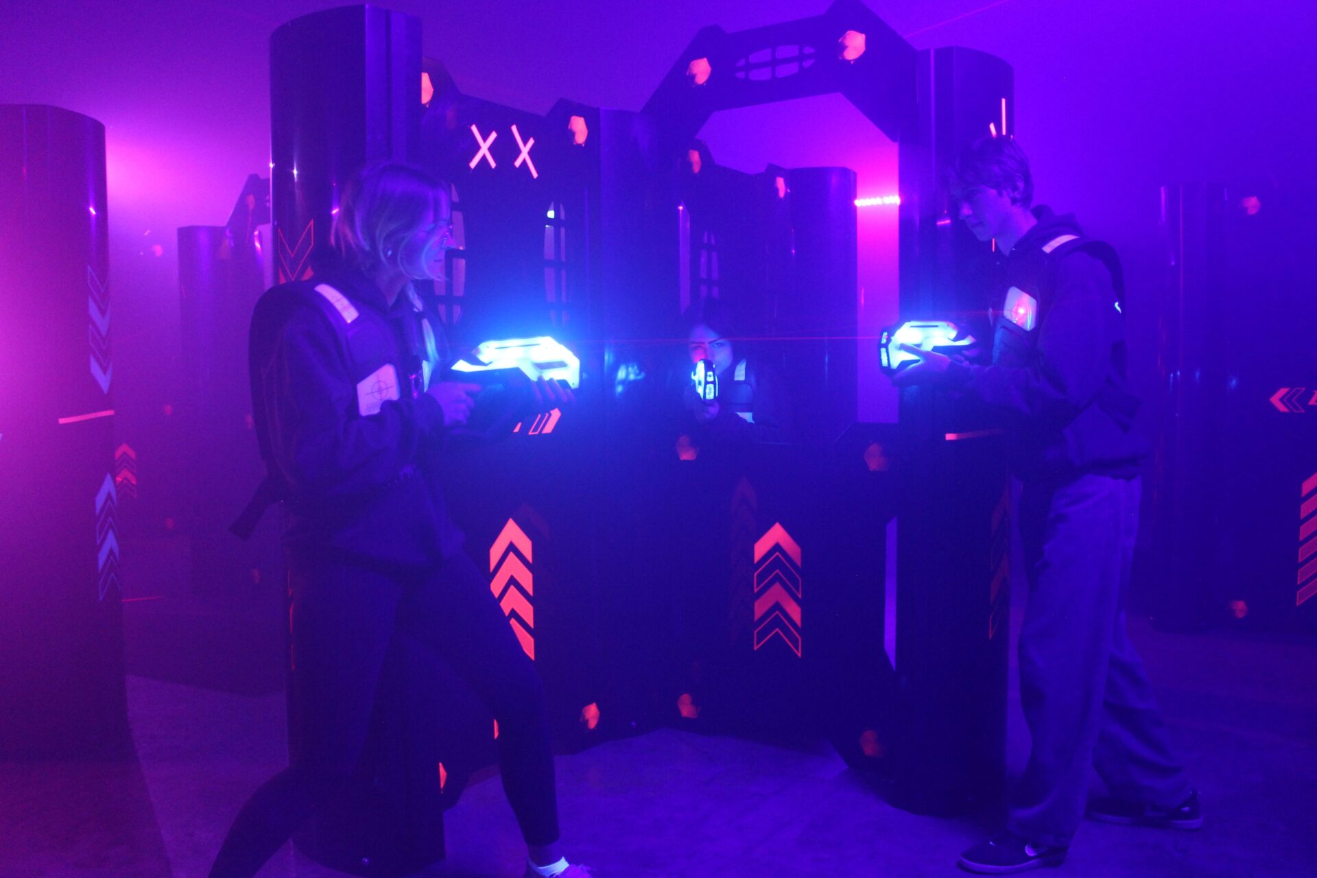 Two teenagers - a boy and a girls - stand facing each other in front of a black wall with fluorescent orange arrows painted on it. The atmosphere is smokey. There are pink, purple, and blue lights colouring the smoke and the faces of the teens. The two teens hold laser tag guns - about the length of their forearms and glowing with blue light. They point their guns at each other.  A third teen - a girl - peeks over an opening in the black wall between the two teens facing off and points her gun at the camera.